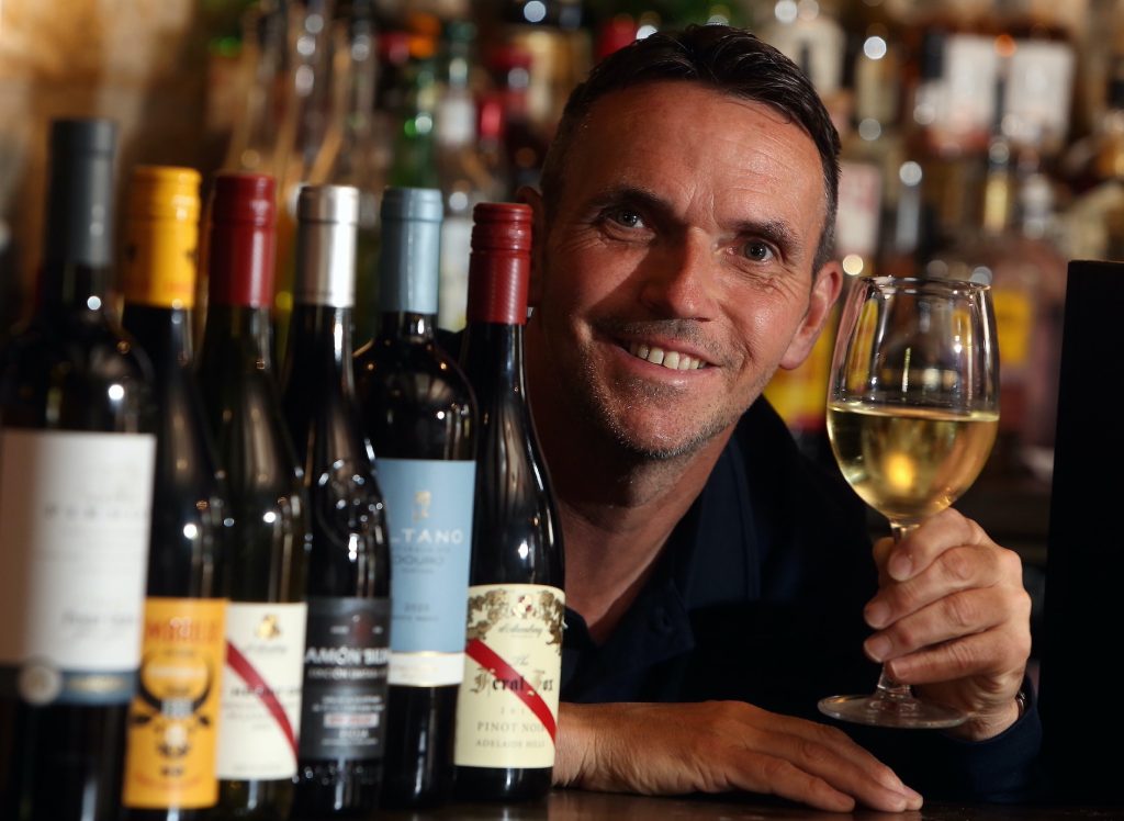 Chris from Cheltenham Local Wine School holding a wine glass with bottles of wine in front of him | StayCotswold